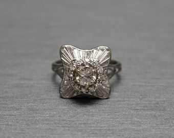 May include: A silver ring with a square-shaped setting featuring a large, clear, round-cut diamond surrounded by smaller diamonds. The ring has a delicate, intricate design.