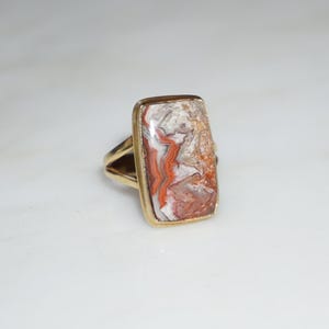 Puede incluir: Anillo dorado con una piedra rectangular con un patrón único de tonos rojos, blancos y marrones. El anillo tiene un diseño de caña dividida. Una pieza llamativa para cualquier ocasión.