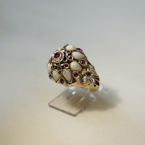 May include: A gold ring with a cluster of white opals and red rubies. The ring is set on a clear plastic ring stand.