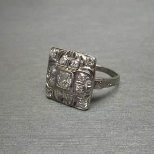 May include: A silver ring with a square-shaped center stone surrounded by smaller diamonds. The ring has a delicate, intricate design.