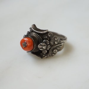 May include: A silver ring with an intricate design featuring a red coral stone.