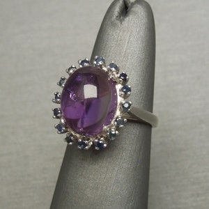 May include: A silver ring featuring a large, oval-shaped, deep purple amethyst gemstone. The amethyst is surrounded by a halo of smaller, dark blue gemstones. The ring is displayed on a gray stand.