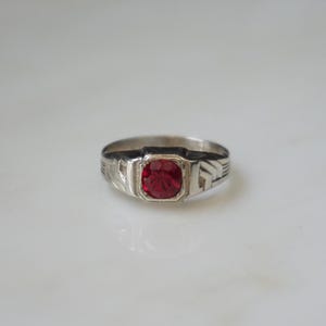May include: A silver-toned ring featuring a square-cut, faceted red stone set in the center. The band displays engraved linear detailing on both sides of the setting, providing a textured appearance to the metal surface surrounding the stone.