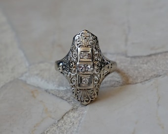 Op de afbeelding: Een zilveren ring met een diamantvormig ontwerp en drie kleine diamanten in het midden.