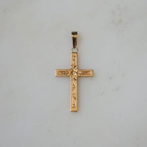 May include: A gold-colored cross pendant with intricate floral and scrollwork detailing. The cross has a loop at the top for a chain. The pendant is displayed against a light-colored background.