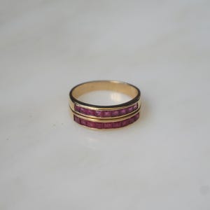 May include: A gold ring with two rows of square-cut, deep red gemstones. The ring has a simple, elegant design, with the gemstones set closely together. The band is a warm gold color, creating a striking contrast with the red gems.