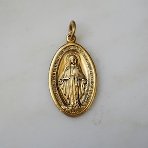May include: Gold-toned oval pendant featuring a detailed depiction of a religious figure with outstretched arms. The pendant has an ornate design with engraved text around the border and a loop for hanging. The pendant is likely made of metal.