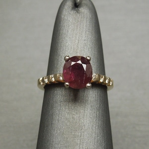 May include: A gold ring with a large, round, red ruby gemstone surrounded by smaller diamonds. The ring is on a gray display stand.