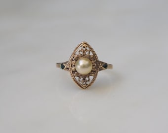 Op de afbeelding: Een vintage goudkleurige ring met een ovaal filigraan ontwerp. De ring heeft een centrale parel en kleine donkergroene accenten aan weerszijden. De ring is geplaatst tegen een witte achtergrond.