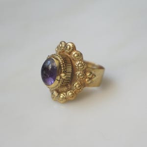 May include: A gold-colored ring with an oval, purple gemstone. The ring has an ornate, textured design around the gemstone and on the band. The ring is set against a white background.