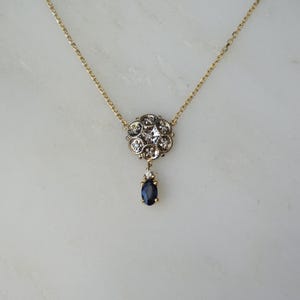 May include: A gold chain necklace with a round pendant featuring a cluster of small diamonds and a single, dangling, dark blue sapphire.
