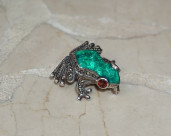 May include: A silver frog brooch with green and red stones. The frog is sitting on a light brown surface.