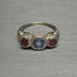May include: A gold ring with three gemstones. The center stone is a blue sapphire, and the two stones on either side are red rubies.