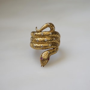 May include: A gold-toned ring shaped like a coiled snake with a textured, scale-like surface. The snake body is accented with small, clear, round stones embedded along its length, and the head features two small, vibrant red stones for eyes.