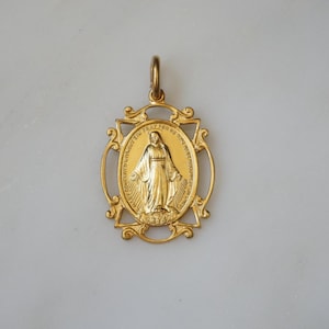 May include: A gold-colored pendant featuring a religious figure within an oval frame. The pendant has an ornate, filigree-style border and a loop for a chain. The background is a light gray.