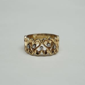 May include: A gold-colored ring with a heart-shaped filigree design. The ring features small, clear gemstones set within the heart shapes. The ring is displayed on a white surface.