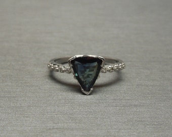 Pode incluir: Um anel de prata com uma gema azul triangular engastada em garras. A banda é decorada com pequenas pedras redondas.