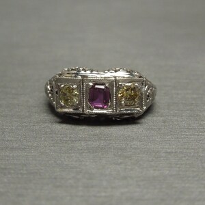 May include: A silver ring with three stones set in a filigree design. The center stone is a rectangular, dark pink stone. The stones on either side are square, yellow stones.