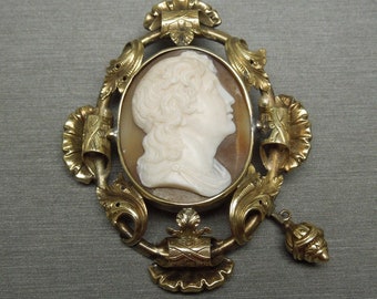 May include: A gold cameo brooch with a white cameo portrait of a person with long hair. The cameo is set in a gold frame with ornate details and a dangling gold ornament.