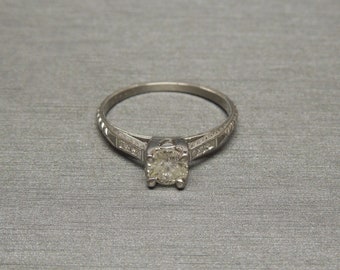 May include: A silver engagement ring with a round, clear center stone and smaller stones on the band.