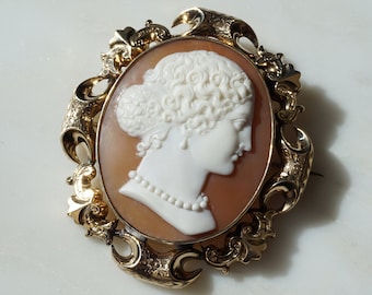 May include: Oval cameo brooch featuring a white profile of a woman with curly hair and a pearl necklace, set against a peach background. The cameo is framed by an ornate gold-colored filigree design.