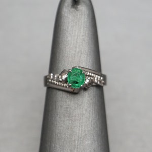 May include: A silver ring with a green emerald gemstone set in a prong setting. The ring has a delicate, intricate design with small diamonds on the band.