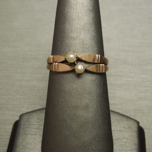 May include: A gold ring with two small pearl stones set in the center. The ring is on a gray display stand.