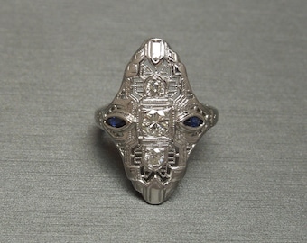 Op de afbeelding: Een zilveren ring met een geometrisch ontwerp met drie diamanten en twee blauwe saffieren.