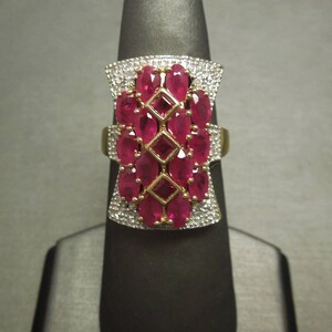 May include: A gold ring with a large red ruby stone set in the center. The ruby is surrounded by smaller diamonds. The ring is on a black display stand.