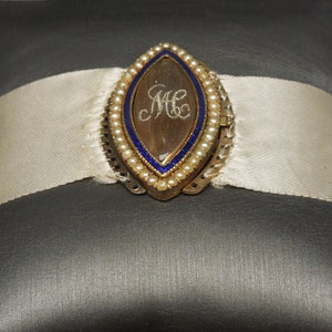 May include: A gold locket with a blue enamel border and a pearl trim. The locket is open and reveals a clear glass insert with the initials "M.C." inscribed inside.