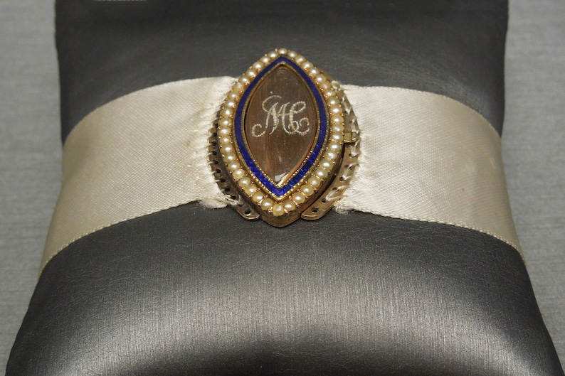 May include: A gold locket with a blue enamel border and a pearl trim. The locket is open and reveals a clear glass insert with the initials "M.C." inscribed inside.