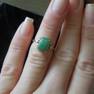 K&ouml;nnte beinhalten: Ein goldener Ring mit einem gr&uuml;nen ovalen Edelstein und kleinen klaren Edelsteinen auf der Ringschiene.
