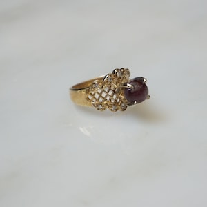 May include: A gold ring with a red gemstone set in a delicate, openwork design. The ring is accented with small, clear stones.