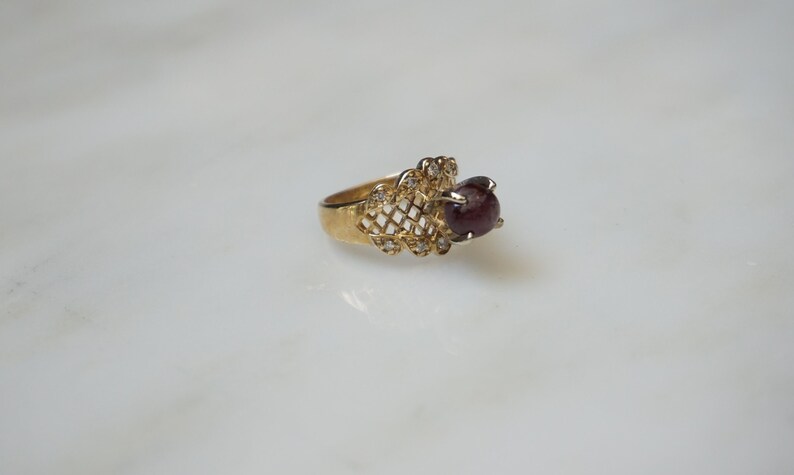 May include: A gold ring with a red gemstone set in a delicate, openwork design. The ring is accented with small, clear stones.
