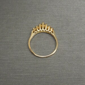 May include: A gold ring with a crown-like design featuring multiple vertical bars. The ring is set against a gray background, showcasing its intricate details and polished finish. The ring's band is smooth and rounded.