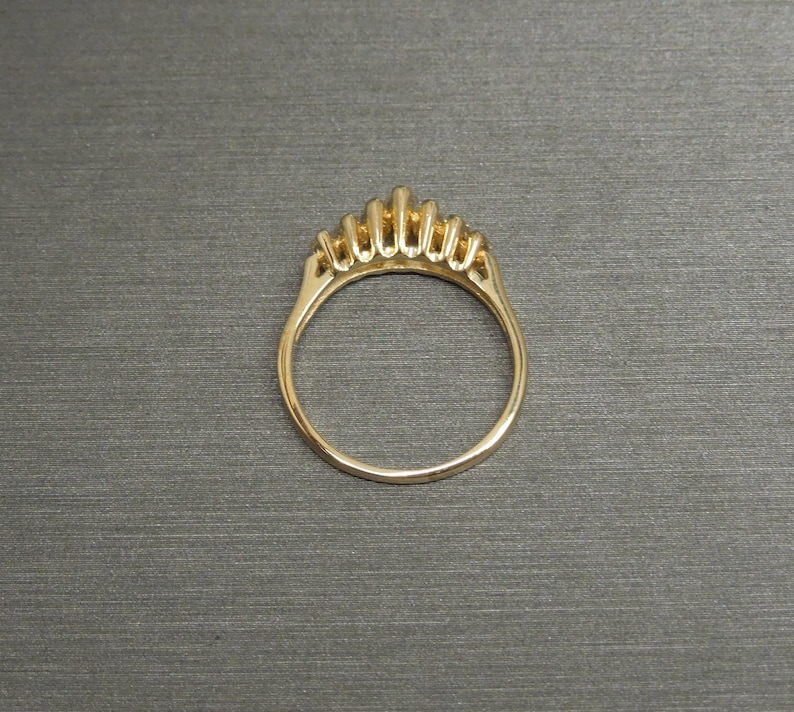 May include: A gold ring with a crown-like design featuring multiple vertical bars. The ring is set against a gray background, showcasing its intricate details and polished finish. The ring's band is smooth and rounded.