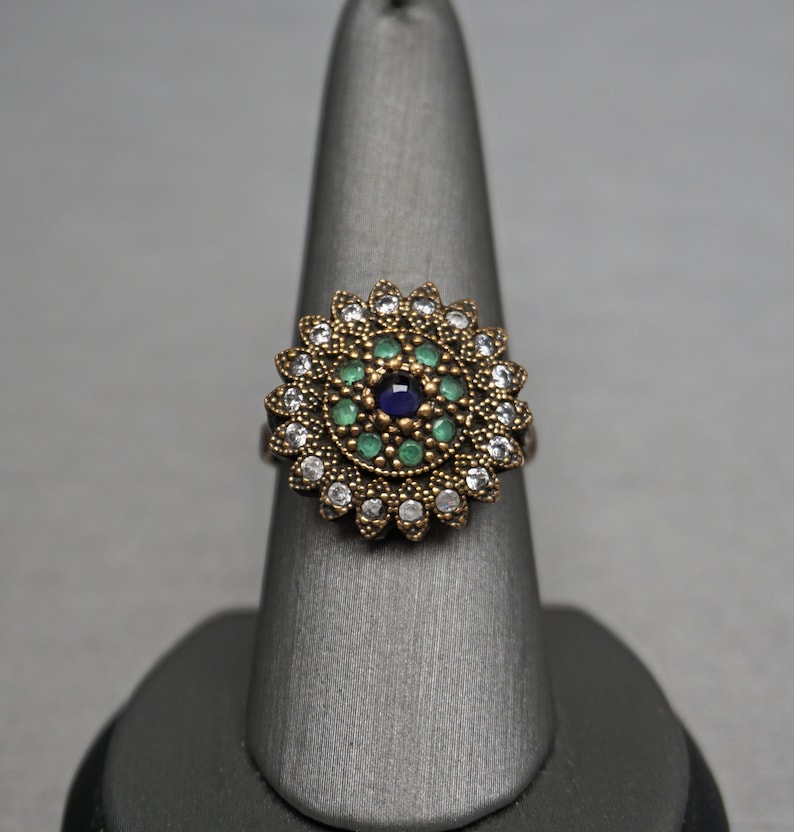 Pu&ograve; includere: Un anello d'oro con un grande designo floreale rotondo. Il centro del designo presenta una pietra preziosa blu circondata da pietre preziose verdi pi&ugrave; piccole. L'anello &egrave; posto su un espositore per anelli grigio.