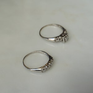 May include: Two silver rings with a row of small, clear gemstones set in the band. The rings are displayed on a white surface, with one ring slightly above the other.