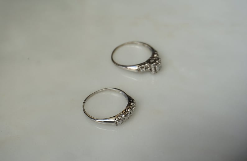 May include: Two silver rings with a row of small, clear gemstones set in the band. The rings are displayed on a white surface, with one ring slightly above the other.