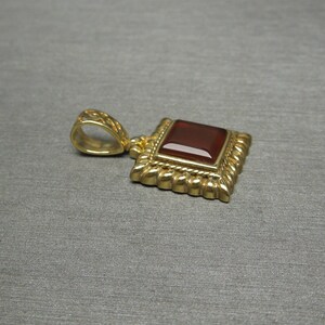 May include: A gold pendant with a square red gemstone set in a decorative frame. The pendant has a loop for attaching to a chain.
