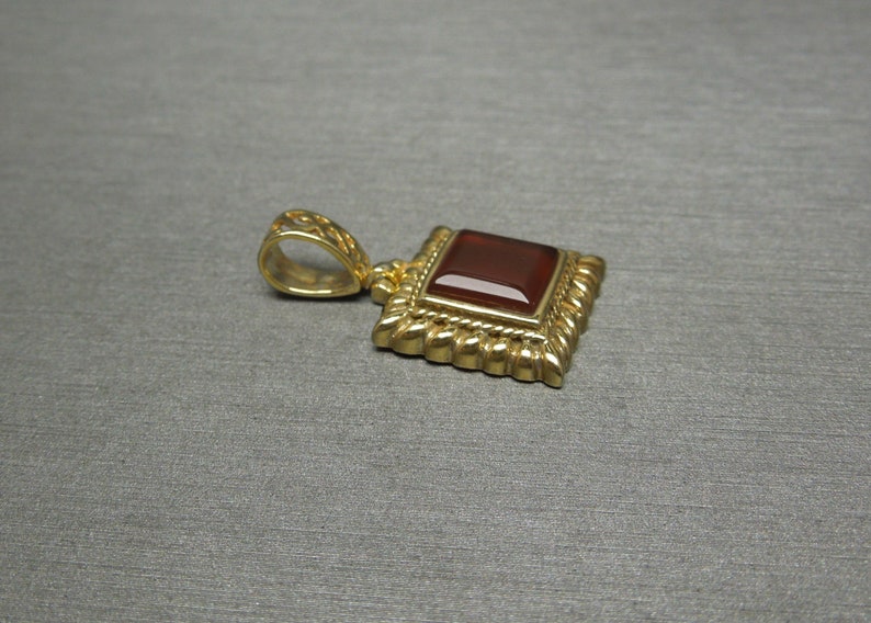 May include: A gold pendant with a square red gemstone set in a decorative frame. The pendant has a loop for attaching to a chain.