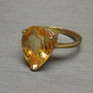 May include: A gold ring featuring a large pear-shaped topaz gemstone. The gemstone is a light orange colour and has a faceted cut.