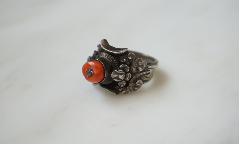 May include: A silver ring with an intricate design featuring a red coral stone.