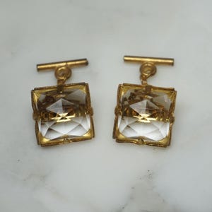 May include: A pair of gold cufflinks with clear crystal accents. The cufflinks have a square shape and a simple design.
