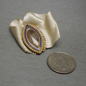 May include: A gold locket with a blue enamel border and a glass insert containing a lock of hair. The locket is decorated with small white pearls.