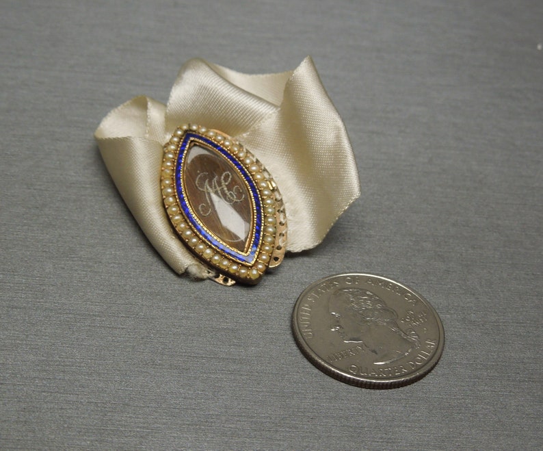 May include: A gold locket with a blue enamel border and a glass insert containing a lock of hair. The locket is decorated with small white pearls.
