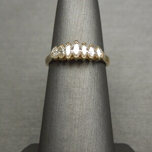 May include: A gold ring featuring a series of diamonds. The ring has a classic design with a row of marquise-cut diamonds and a round diamond. The ring is displayed on a gray stand.