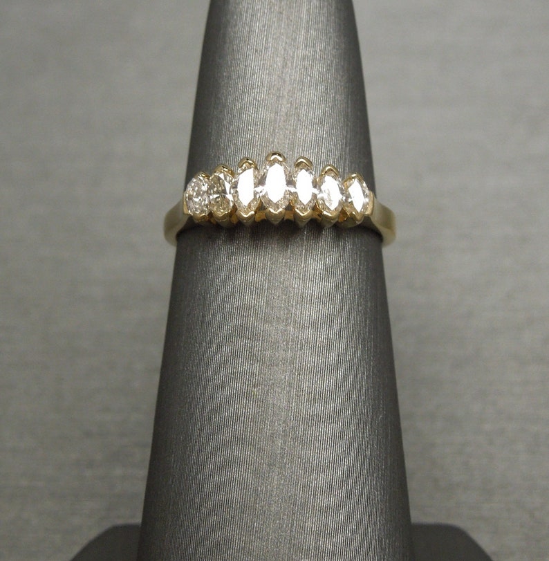 May include: A gold ring featuring a series of diamonds. The ring has a classic design with a row of marquise-cut diamonds and a round diamond. The ring is displayed on a gray stand.