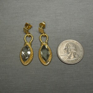 May include: A pair of gold earrings with a teardrop-shaped grey gemstone. The earrings have a twisted rope design and a small loop at the top.