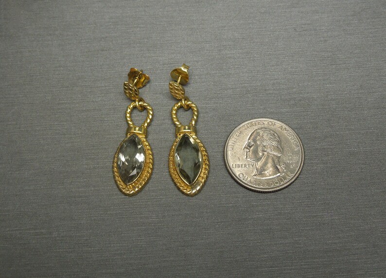 May include: A pair of gold earrings with a teardrop-shaped grey gemstone. The earrings have a twisted rope design and a small loop at the top.